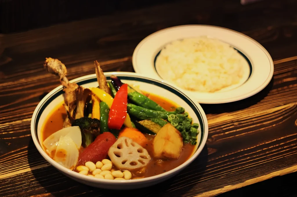 A bowl of vibrant vegetable curry features mushrooms, peppers, potatoes, lotus root, and beans in rich broth beside a plate of fluffy rice on a wooden table.