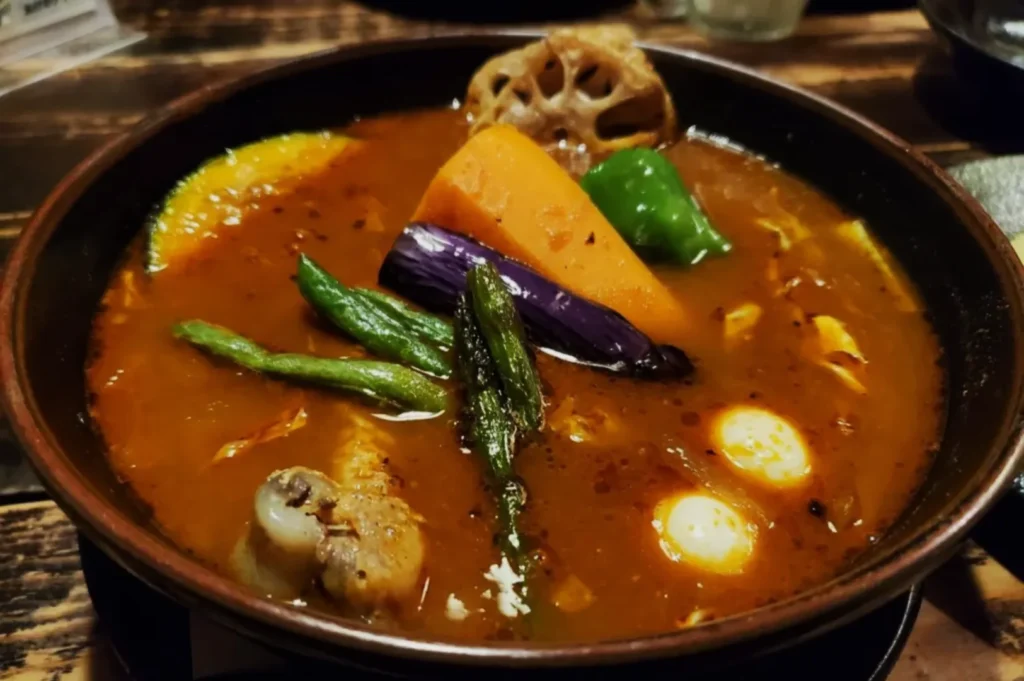 A rich stew with vibrant vegetables like carrot, eggplant, and beans in a brown broth. The dish's warmth and variety evoke comfort and heartiness.
