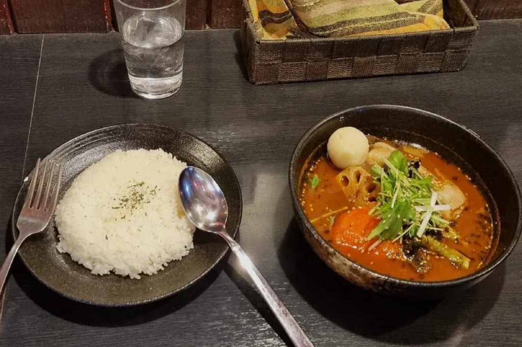 A bowl of vibrant curry with vegetables and an egg sits next to a plate of white rice garnished with herbs. A water glass and utensils accompany.