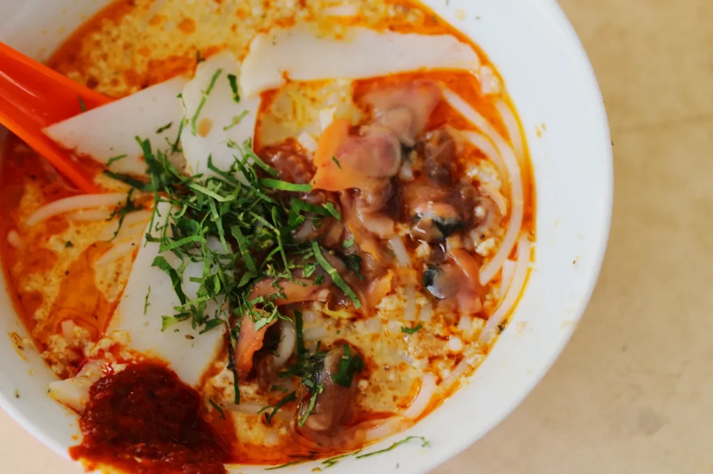 Creamy laksa in a white bowl, topped with fresh herbs, sliced fish cake, vibrant orange broth, and a dollop of chili paste. A vibrant, aromatic dish.