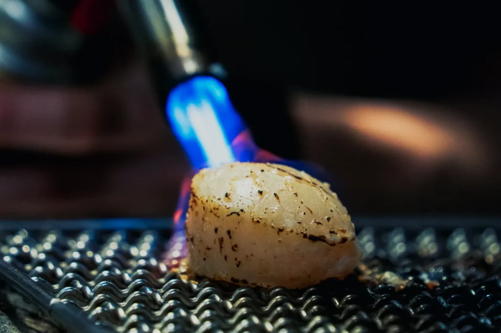 A scallop being seared on a metal grill with a blue flame torch. The scallop is lightly charred, highlighting a chef's precise cooking technique.