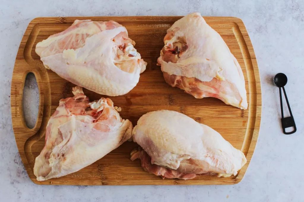 Four raw chicken breasts on a wooden cutting board. A measuring spoon is placed on the right, contrasting with the stone-like surface underneath.