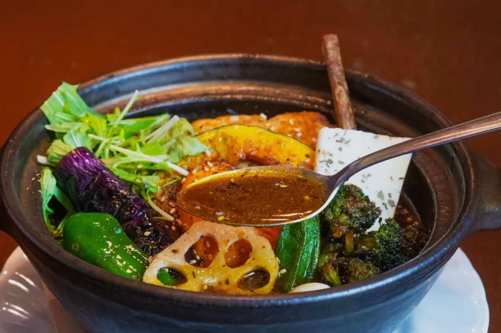 A rich vegetable curry in a black pot with a spoonful of broth, showing lotus root, tofu, and colorful veggies, evoking warmth and savoriness.
