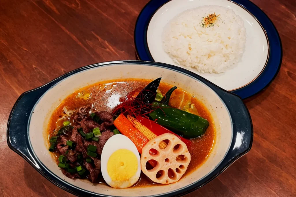 A bowl of vibrant vegetable curry with sliced beef, boiled egg, lotus root, and green onions. Served beside a plate of white rice on a wooden table.