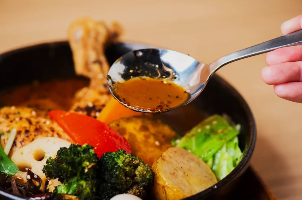 A close-up of a spoon being held over a vibrant vegetable curry. Bright vegetables like broccoli, red pepper, and cabbage surround a rich amber broth.