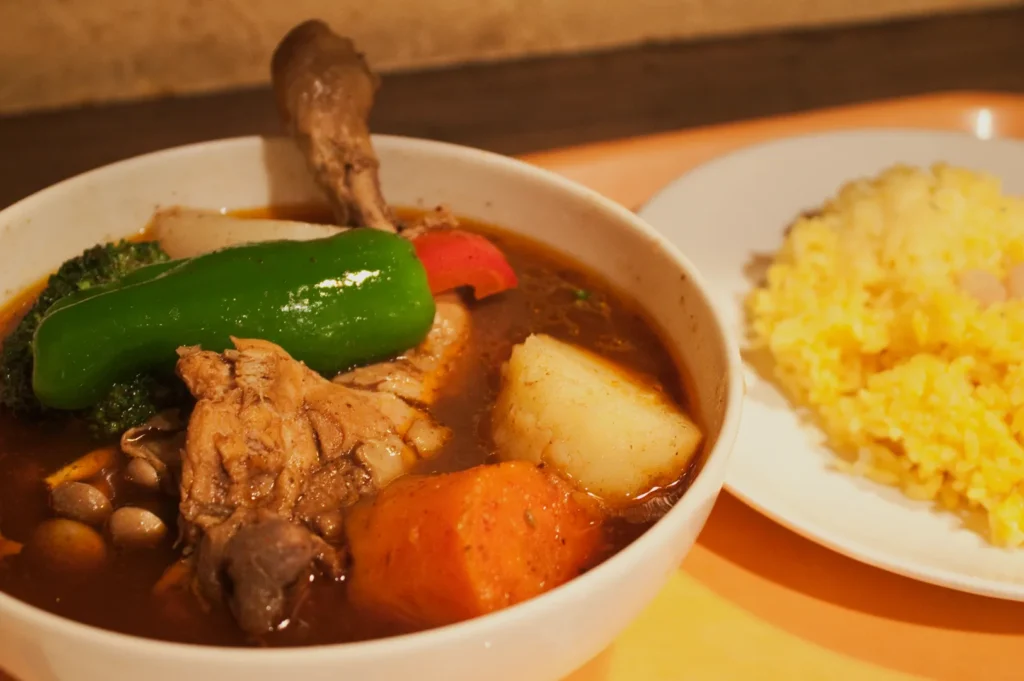 A bowl of rich, colorful stew with chicken, bell pepper, potato, broccoli, and carrot is paired with a plate of yellow rice, conveying warmth and comfort.