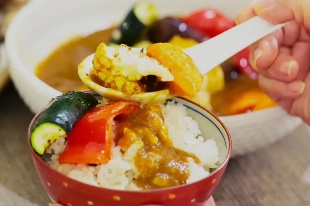 A bowl of rice with golden curry topped with zucchini, red bell pepper, and eggplant. A spoon scoops curry from the bowl, suggesting a comforting meal.