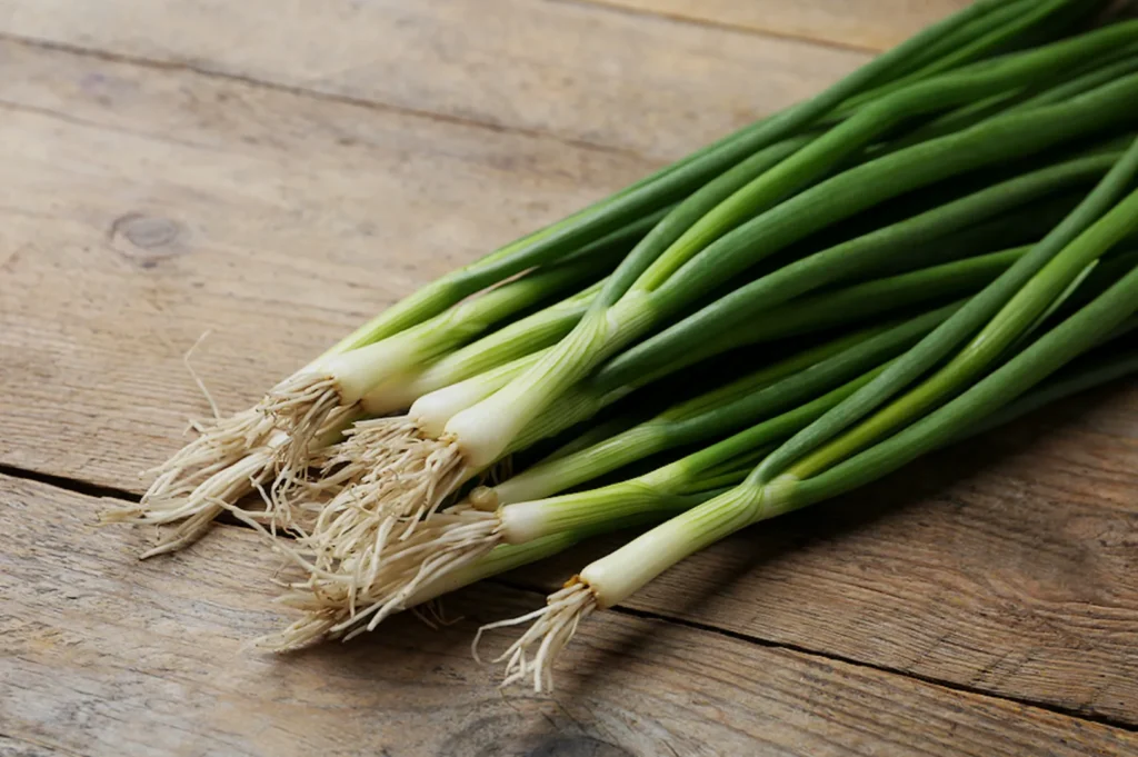 A bunch of fresh green onions lies on a rustic wooden table. The vibrant green stalks and white bulbs convey freshness and a natural, earthy tone.