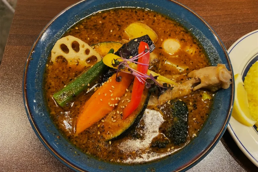 A blue bowl of vibrant curry soup with a chicken drumstick, assorted vegetables, and sesame seeds, evoking a warm, savory aroma.