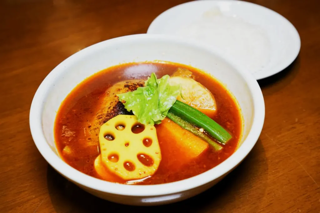 A bowl of vibrant red soup curry with lotus root, carrot, and green okra on a wooden table, accompanied by a small plate of white rice.