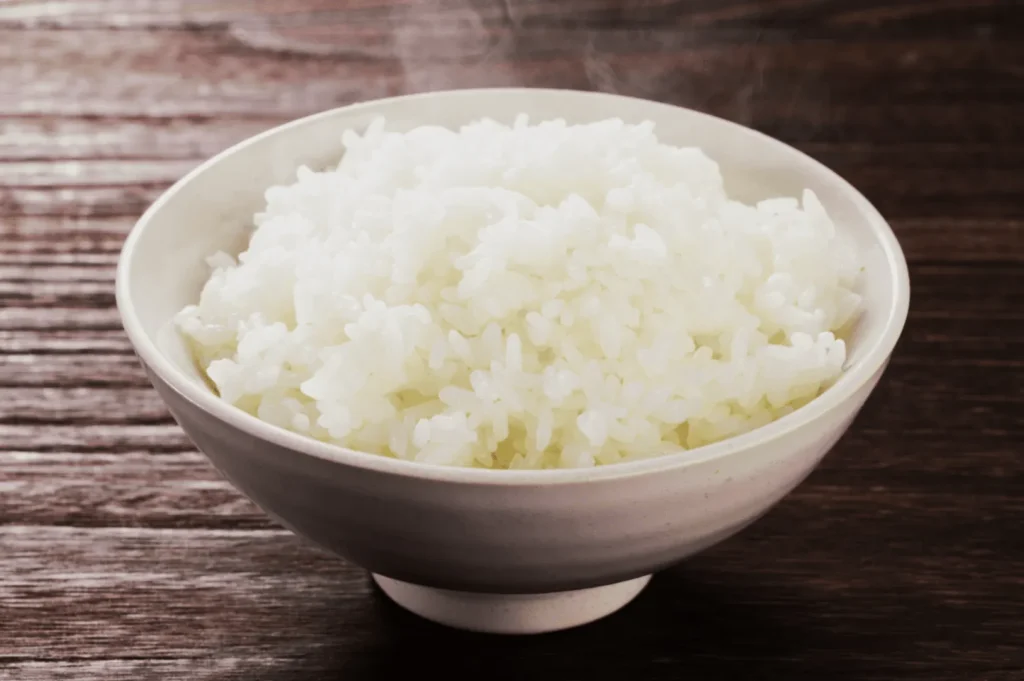 A steaming bowl of fluffy white rice sits on a wooden table, conveying warmth and simplicity. The neutral tones create a cozy and inviting atmosphere.