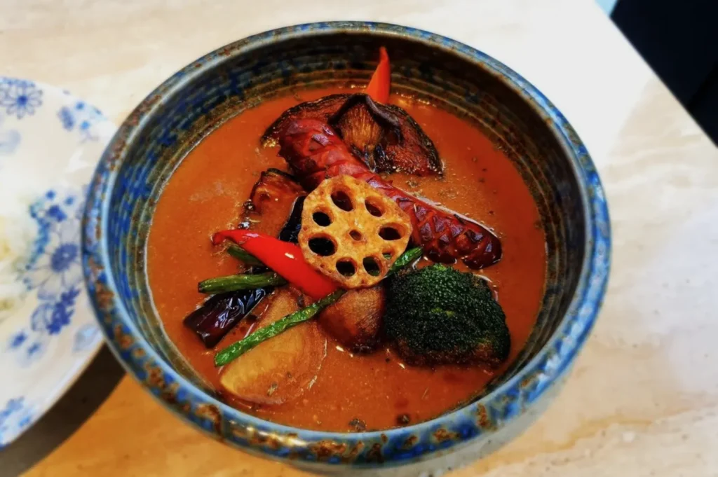 A vibrant curry bowl with a rich orange broth, featuring lotus root, broccoli, red peppers, and sausages, evokes warmth and spice.