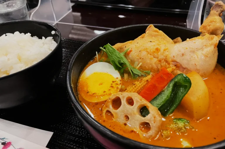 A bowl of curry soup with chicken, boiled egg, lotus root, and vegetables next to a bowl of white rice, set on a dark wooden table.