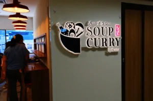 A sign for "Soup Curry" with a graphic of a bowl and various ingredients, located on a wall in a restaurant setting.