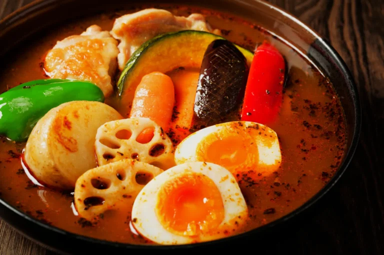A vibrant bowl of Japanese curry soup features colorful vegetables, soft-boiled eggs, and chicken. The rich broth looks spicy and inviting.