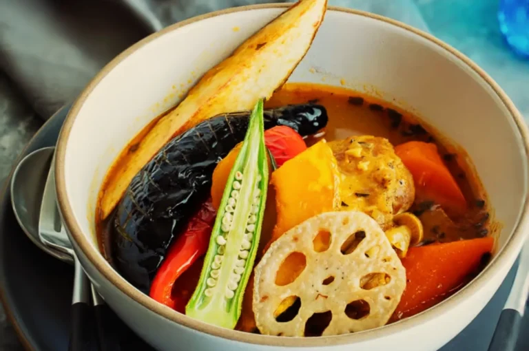 A bowl of vibrant curry soup with grilled eggplant, yellow and red bell peppers, okra, chicken, lotus root, and a potato wedge on top.