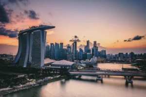 Skyline of Singapore at sunset with Marina Bay Sands and city skyscrapers. The sky is a mix of orange, purple, and pink hues. Calm, serene atmosphere.