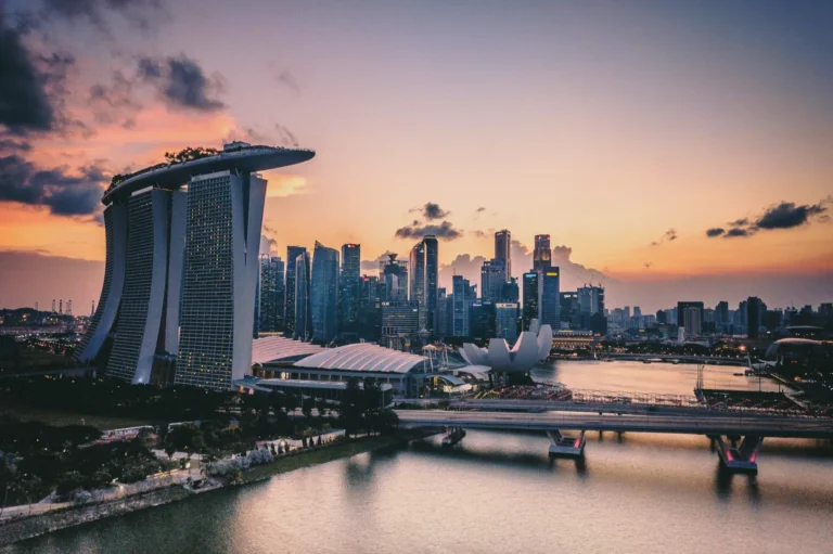 Skyline of Singapore at sunset with Marina Bay Sands and city skyscrapers. The sky is a mix of orange, purple, and pink hues. Calm, serene atmosphere.