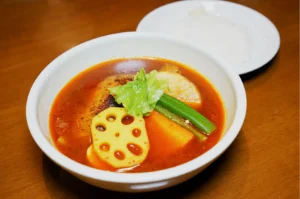 A bowl of vibrant red soup with sliced lotus root, okra, carrot, celery, and a dumpling, on a wooden table. A plate of rice is beside the bowl.