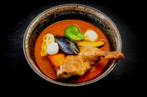 A rustic bowl of rich red curry features a crispy chicken leg, vibrant bell peppers, carrot, a black mussel, boiled eggs, and lotus root slices.