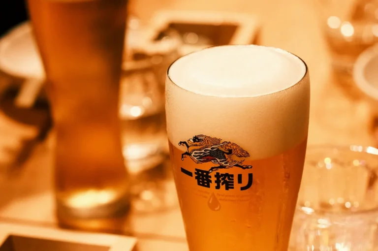 A frosty glass of beer with a creamy head sits on a warmly lit table, surrounded by blurred glassware, creating a cozy and inviting atmosphere.