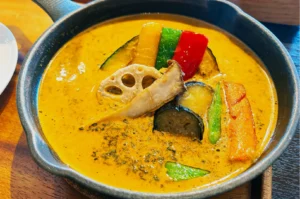 A black bowl filled with vibrant orange curry, topped with colorful vegetables including bell peppers, lotus root, and squash slices. The dish looks warm and inviting.