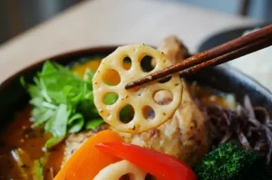 Close-up of chopsticks holding a slice of lotus root over a colorful bowl of curry with broccoli, red peppers, and greens, conveying a fresh, vibrant meal.