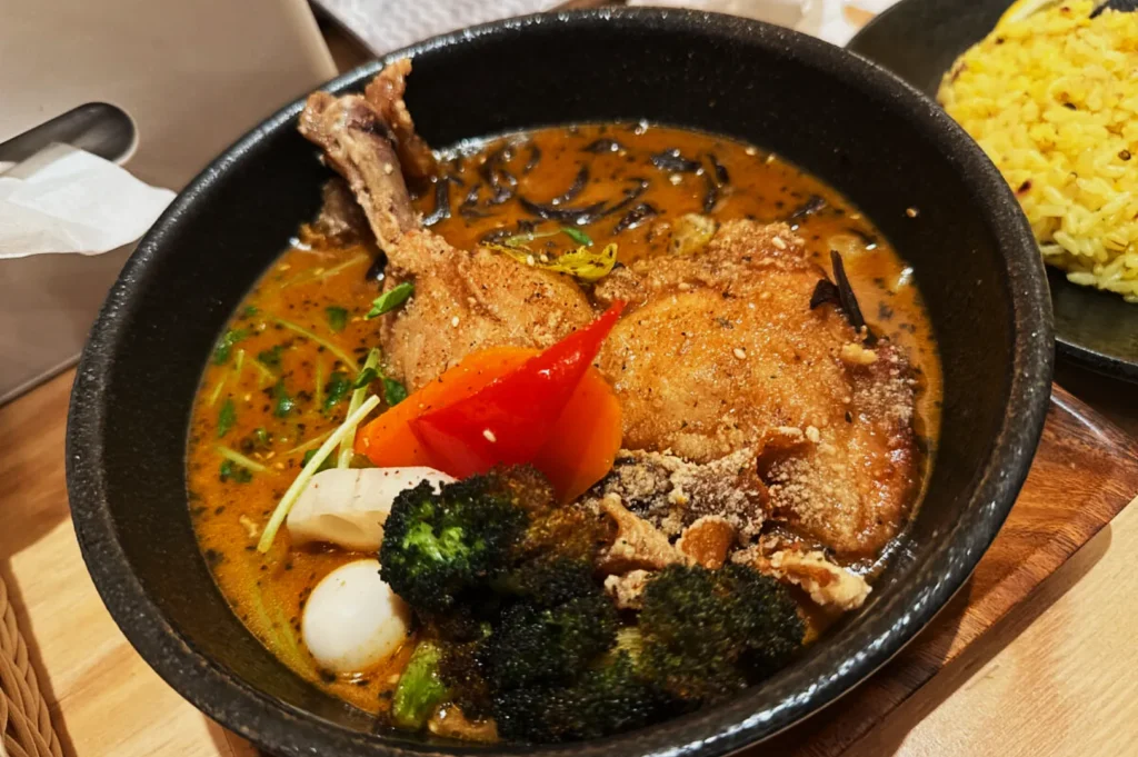 A black bowl of curry with chicken leg, vibrant red pepper, broccoli, quail eggs, and other vegetables. Adjacent is a plate of yellow rice.