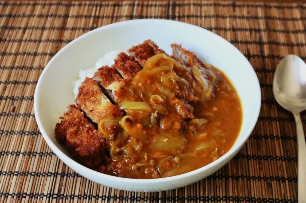 A white bowl of Japanese curry with breaded cutlet and rice on a bamboo mat. A spoon lies nearby, evoking a warm, hearty meal.
