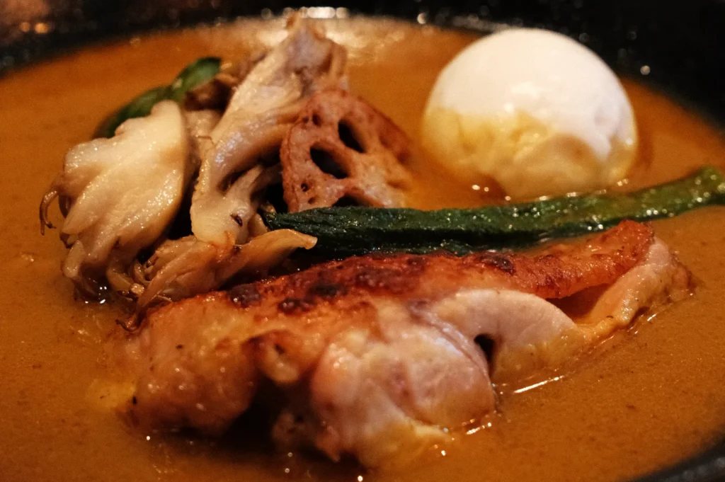 A bowl of curry featuring chicken, mushrooms, lotus root, green vegetables, and a poached egg.