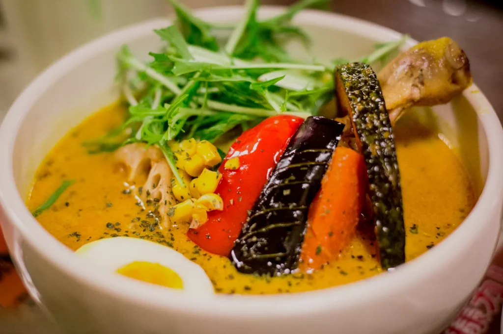 A bowl of Japanese curry soup with chicken leg, bell pepper, eggplant, carrot, corn, lotus root, boiled egg, and fresh greens, creating a vibrant, hearty dish.