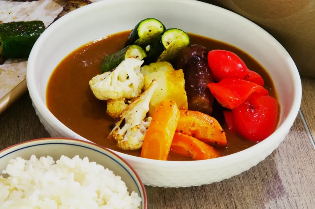 A bowl of vegetable curry featuring zucchini, cauliflower, bell peppers, potatoes, and carrots in rich sauce. Served beside a bowl of white rice.
