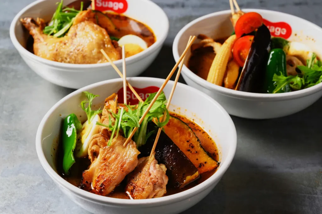 Three bowls of Japanese curry soup are arranged, featuring grilled vegetables, skewered meats, and fresh greens, evoking a warm, savory ambiance.
