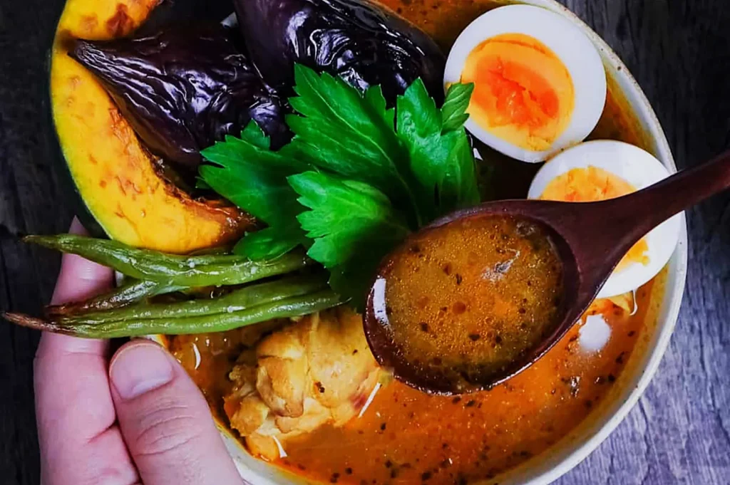A vibrant bowl of curry with colorful toppings: grilled eggplant, squash, green beans, half-sliced boiled eggs, and leafy greens, held by a hand. A wooden spoon rests on top, conveying warmth and flavor.