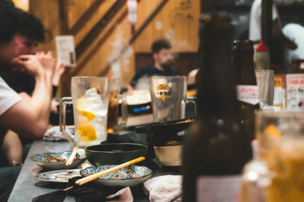 A cozy restaurant scene with people chatting at a table cluttered with plates, chopsticks, and drink glasses. Warm lighting creates a lively, intimate atmosphere.