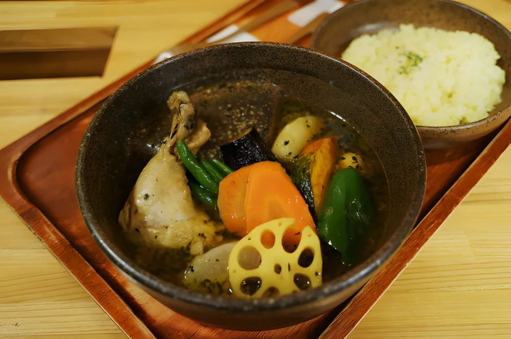 A hearty bowl of soup with chicken, carrots, green beans, eggplant, green bell pepper, and lotus root served alongside a bowl of rice on a wooden tray.