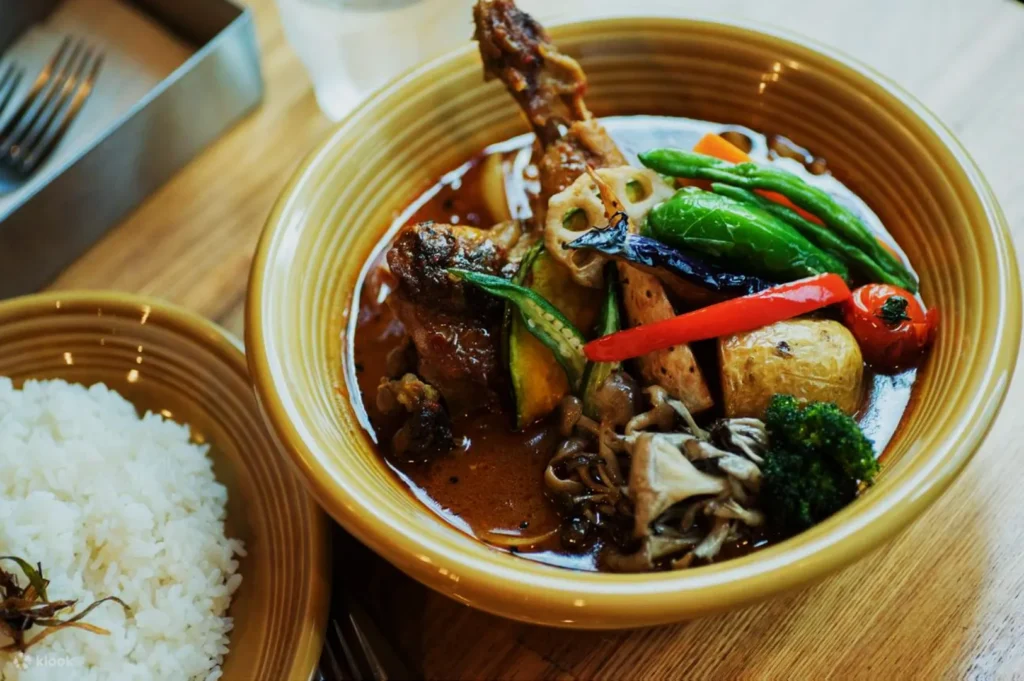 A bowl of flavorful curry soup with chicken, assorted vegetables, and garnishes, alongside a bowl of fluffy white rice on a wooden table.