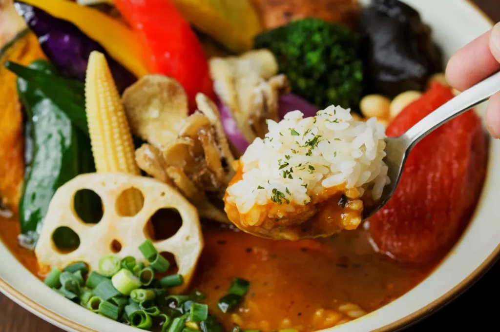 A vibrant vegetable curry plate with rice on a spoon. Features lotus root, baby corn, peppers, mushrooms, and green onion, evoking freshness and flavor.