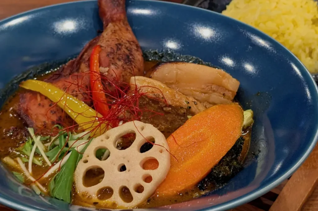 A vibrant bowl of Japanese curry soup features lotus root, carrot, colorful peppers, and seasoned chicken. Served with bright yellow rice on the side.