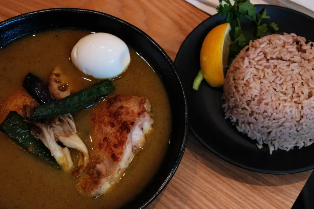 A bowl of curry with chicken, egg, and assorted vegetables, alongside a plate of rice and a slice of lemon.