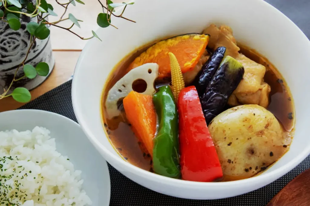 A vibrant bowl of Japanese curry soup features colorful vegetables like pumpkin, eggplant, and peppers. A side of steamed rice with herbs accompanies it.