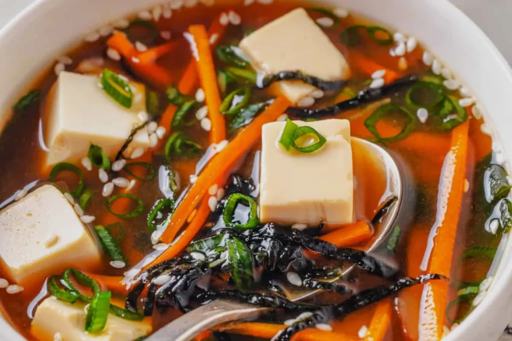 A bowl of miso soup with tofu cubes, carrot strips, seaweed, chopped green onions, and sesame seeds, creating a fresh and inviting appearance.