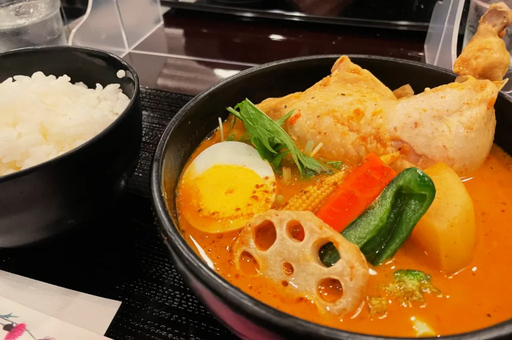 A bowl of spicy curry soup with chicken, half a boiled egg, vegetables, and lotus root. Served with a bowl of white rice. Cozy and appetizing atmosphere.