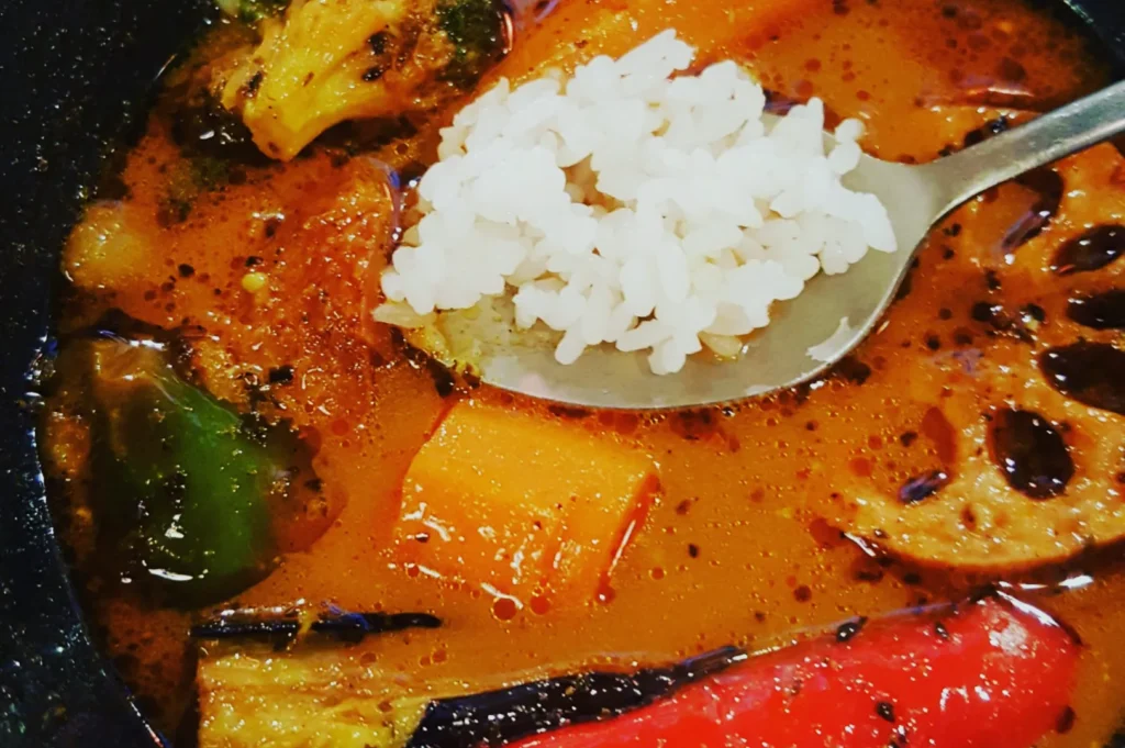 A colorful bowl of Japanese curry soup with vegetables like broccoli, lotus root, and carrots. A spoon holds a mound of white rice. Warm and inviting.