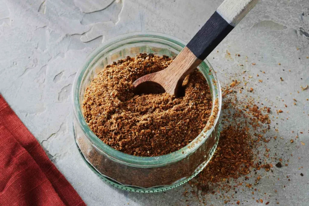 A glass container filled with brown spice powder is shown on a textured grey surface. A wooden spoon rests inside. A red cloth lies on the left.