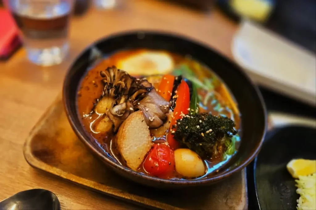 A colorful bowl of soup with assorted vegetables, including mushrooms, carrots, cherry tomatoes, and broccoli, garnished with sesame seeds. Warm, inviting meal setting.