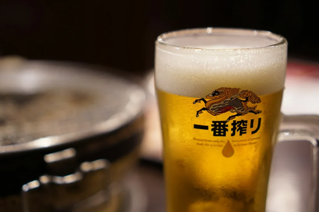 A frosty mug of golden beer with a frothy head, featuring Japanese text and a mythical creature graphic. Blurred background suggests a cozy atmosphere.
