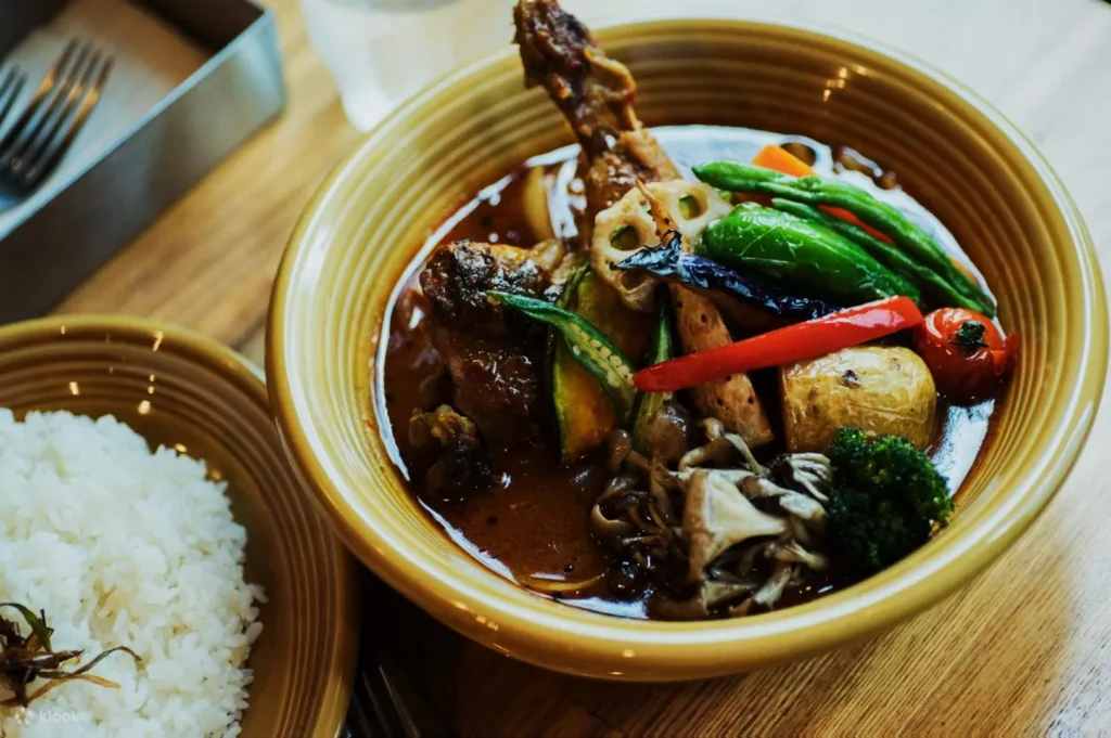 A hearty bowl of rich soup curry filled with assorted vegetables, chicken, and mushrooms. A side of white rice sits nearby on the wooden table.