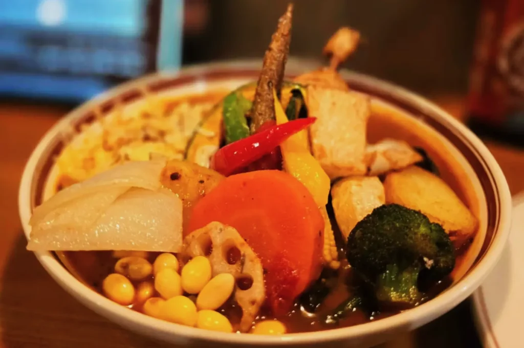 A vibrant bowl of Japanese curry soup is filled with colorful vegetables, tofu, and a chili pepper, creating a warm, inviting, and hearty appearance.