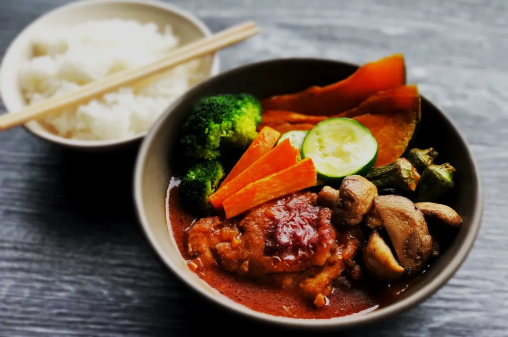 A bowl of vibrant curry with chicken, broccoli, carrots, cucumber, mushrooms, and squash. Steamed white rice with chopsticks is beside it.